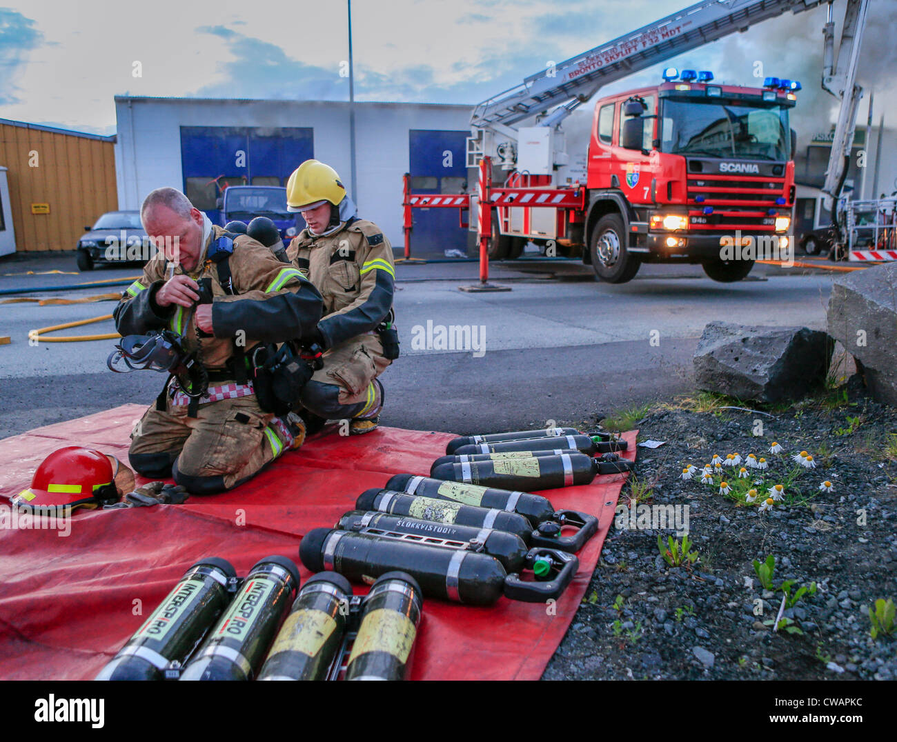 Firefighter assisting team member with oxygen tanks. Auto repair shop on fire in suburb of Reykjavik, Iceland Stock Photo