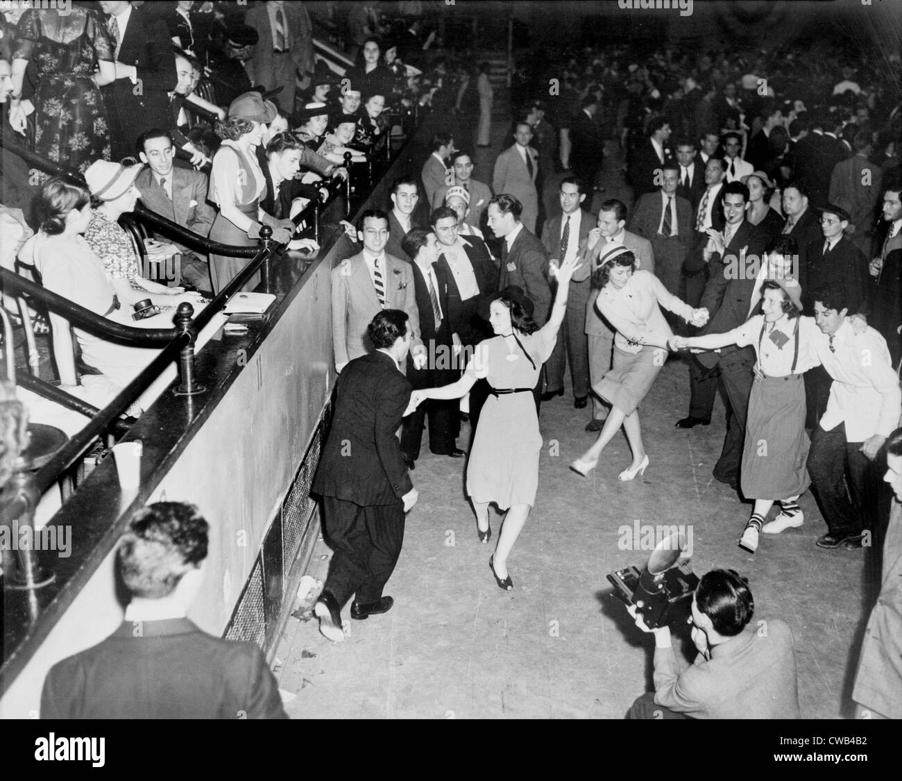 Couples jitterbug dancing on a dance floor, with a photographer in the foreground and the audience in raised seating on the Stock Photo