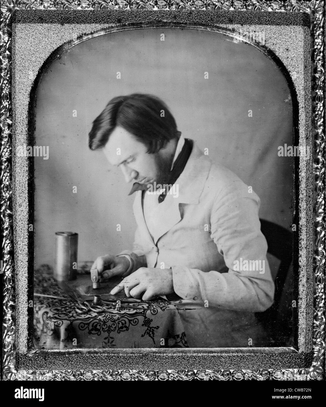 Occupational portrait of a tinworker seated at table with tablecloth, working with a mallet, snips, compass, and metal Stock Photo