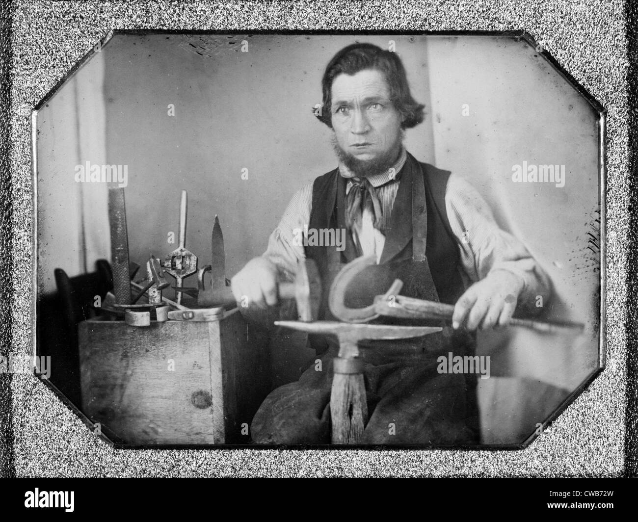 Occupational portrait of a blacksmith, working on a horseshoe at an anvil, other tools to his side. quarter plate Stock Photo