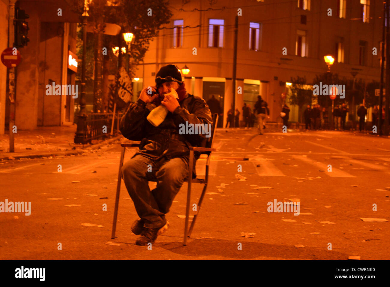 A man rests on a burnt chair in the middle of the street during a night of violence after a demonstration in Athens. Stock Photo