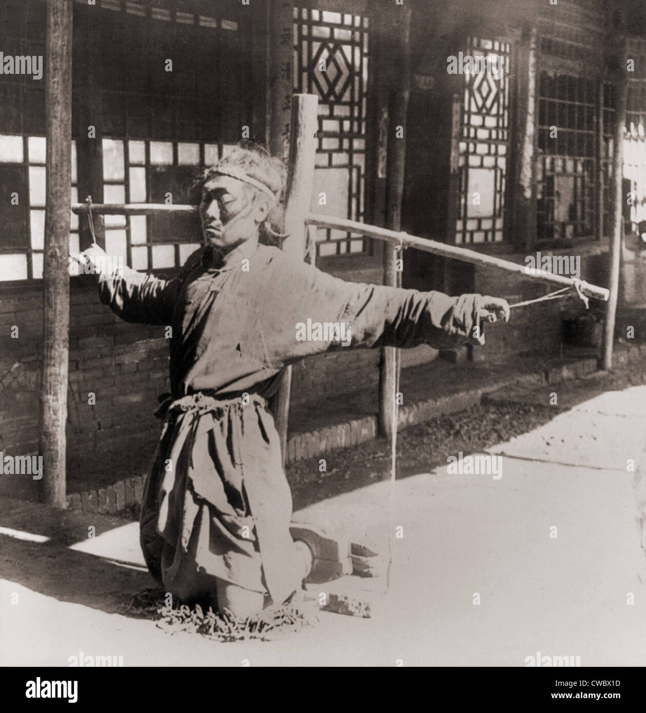 Chinese prisoner kneeling on chains, with his ankles in stocks, while his arms are tied by the