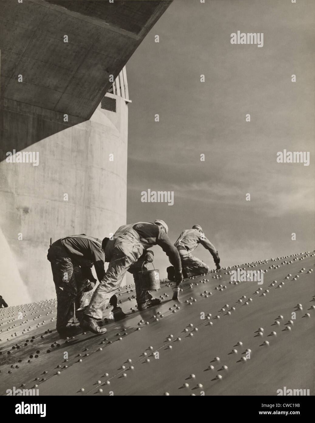 Three workers painting on a slanted wall of riveted-steel plates on the Hoover Dam spillway. Ca. 1940. Stock Photo