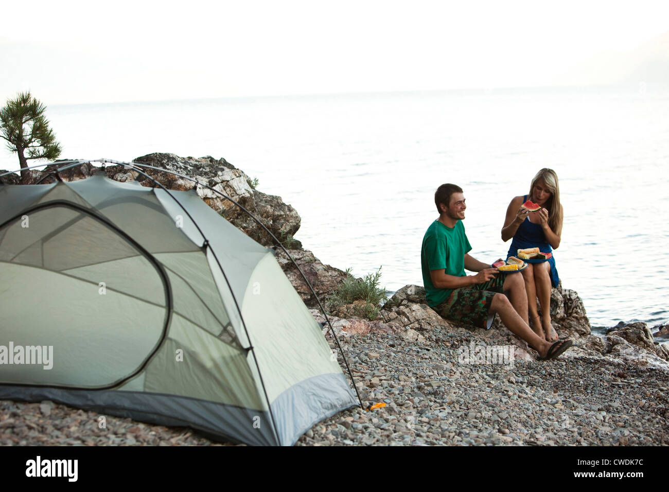 Two young adults camping smile while eating dinner next to a lake in Idaho. Stock Photo