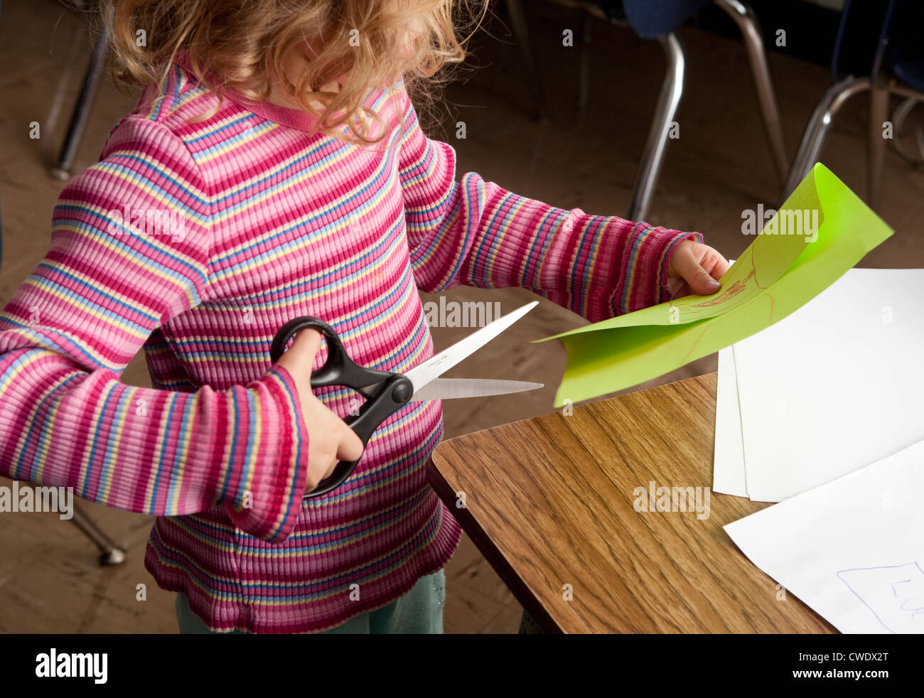 young white 5 year old girl in colorful sweater, uses large dangerous scissors to cut through green paper without supervision Stock Photo