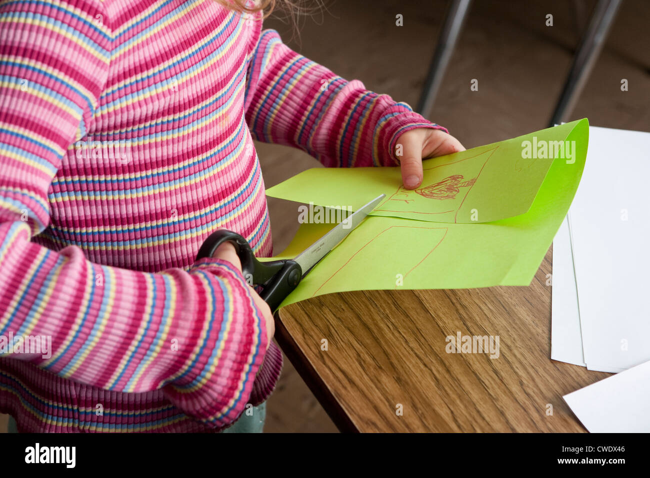 young white 5 year old girl in colorful sweater, uses large dangerous scissors to cut through green paper without supervision Stock Photo