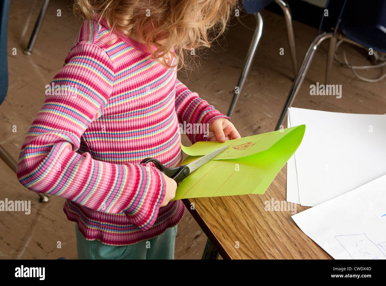 young white 5 year old girl in colorful sweater, uses large dangerous scissors to cut through green paper without supervision Stock Photo