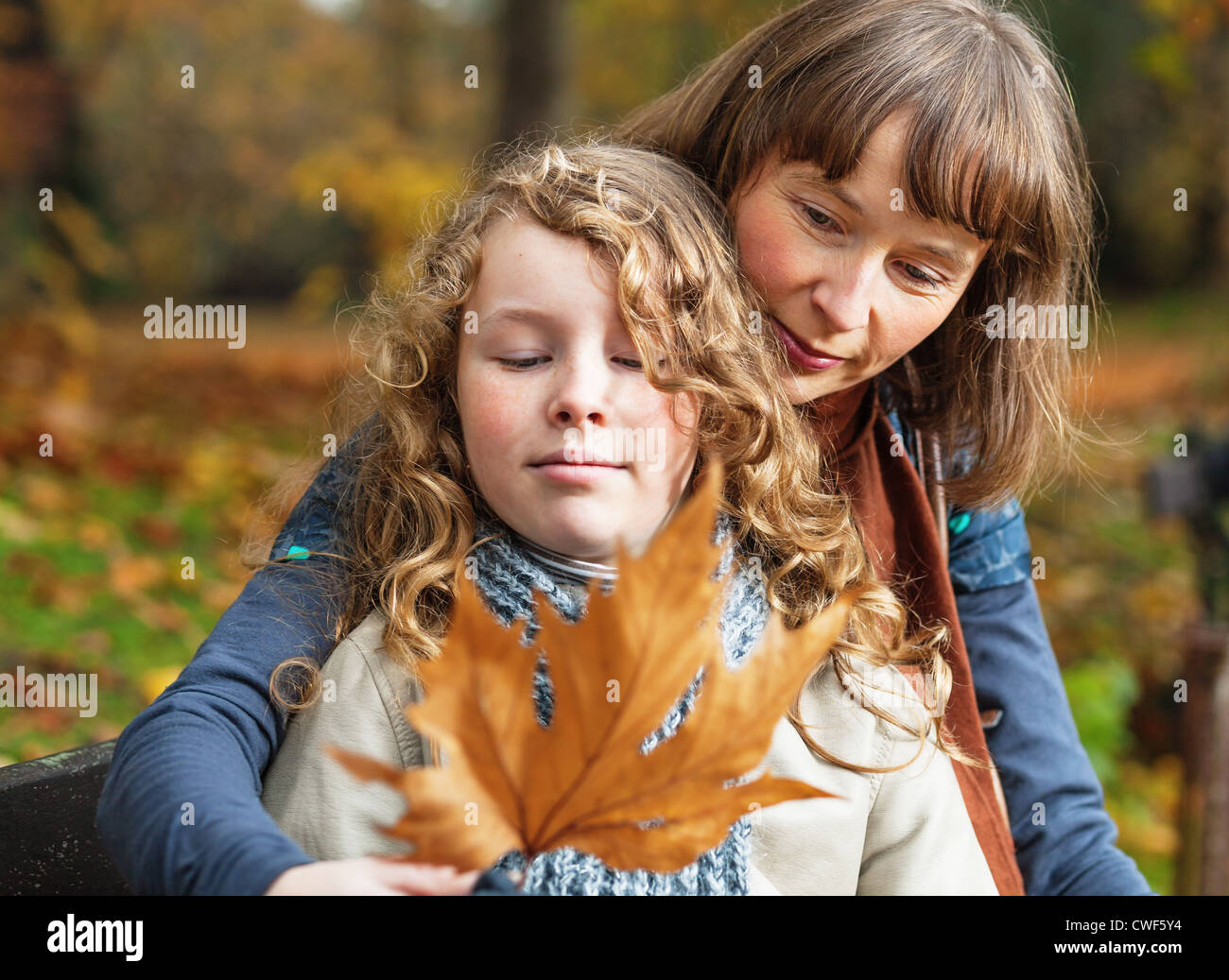 Mother and her teenager daughter sitting on a bench in an autumn park and looking at fallen leaf Stock Photo
