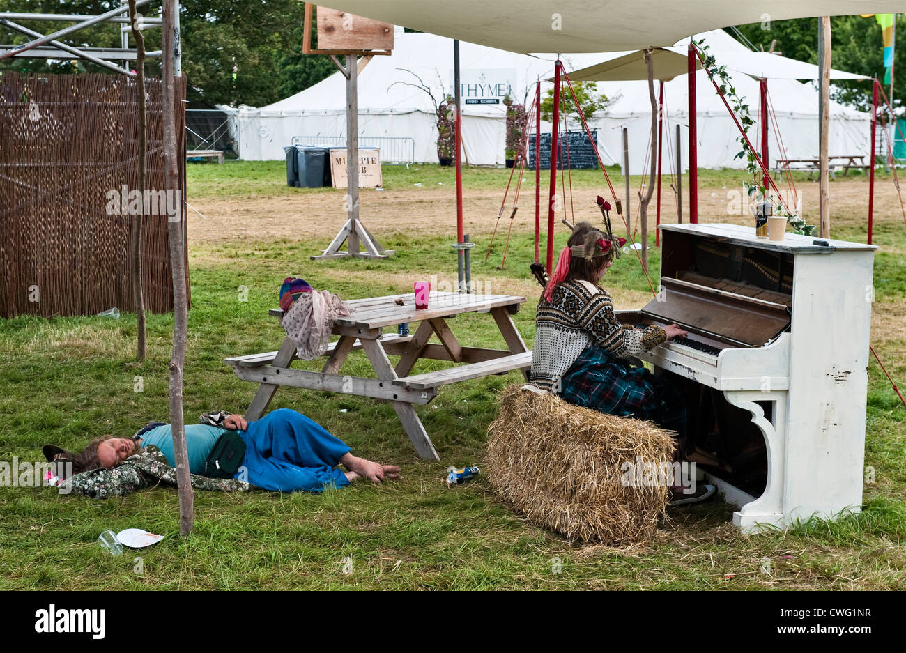 A festival goer asleep on the ground on the morning after Wilderness music festival, Cornbury, Oxfordshire, UK, while another quietly plays a piano Stock Photo