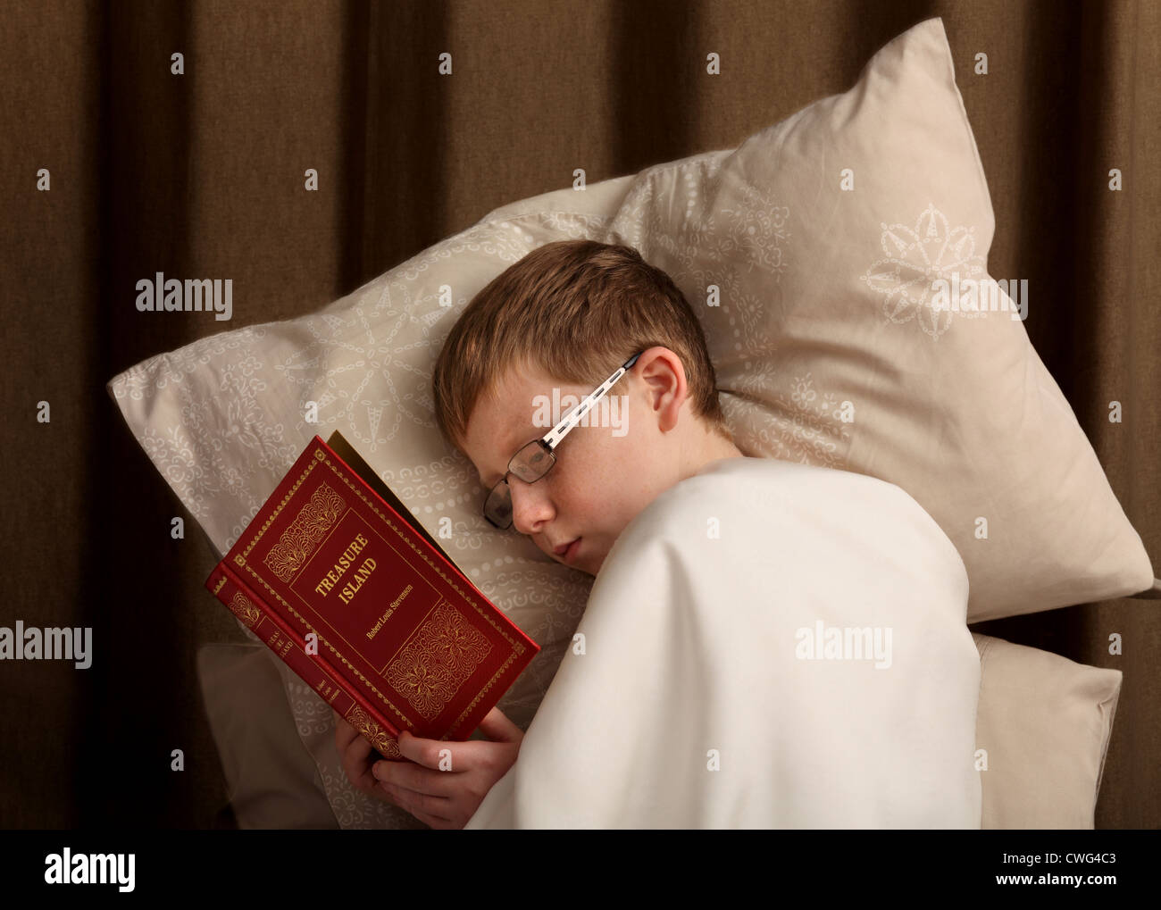A  boy 12 to 13 years old boy, laying on pillows with a sheet over him while reading 'Treasure Island'. The book is not my copyright Stock Photo