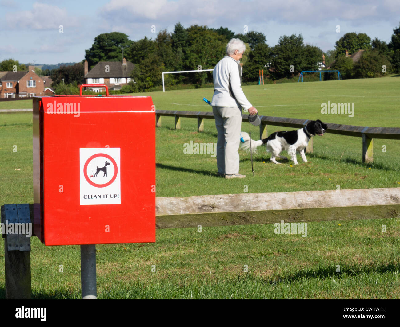 Dog waste bin by playing fields in an urban park public space with a woman walking a dog on daily walk in background. England UK Britain Stock Photo