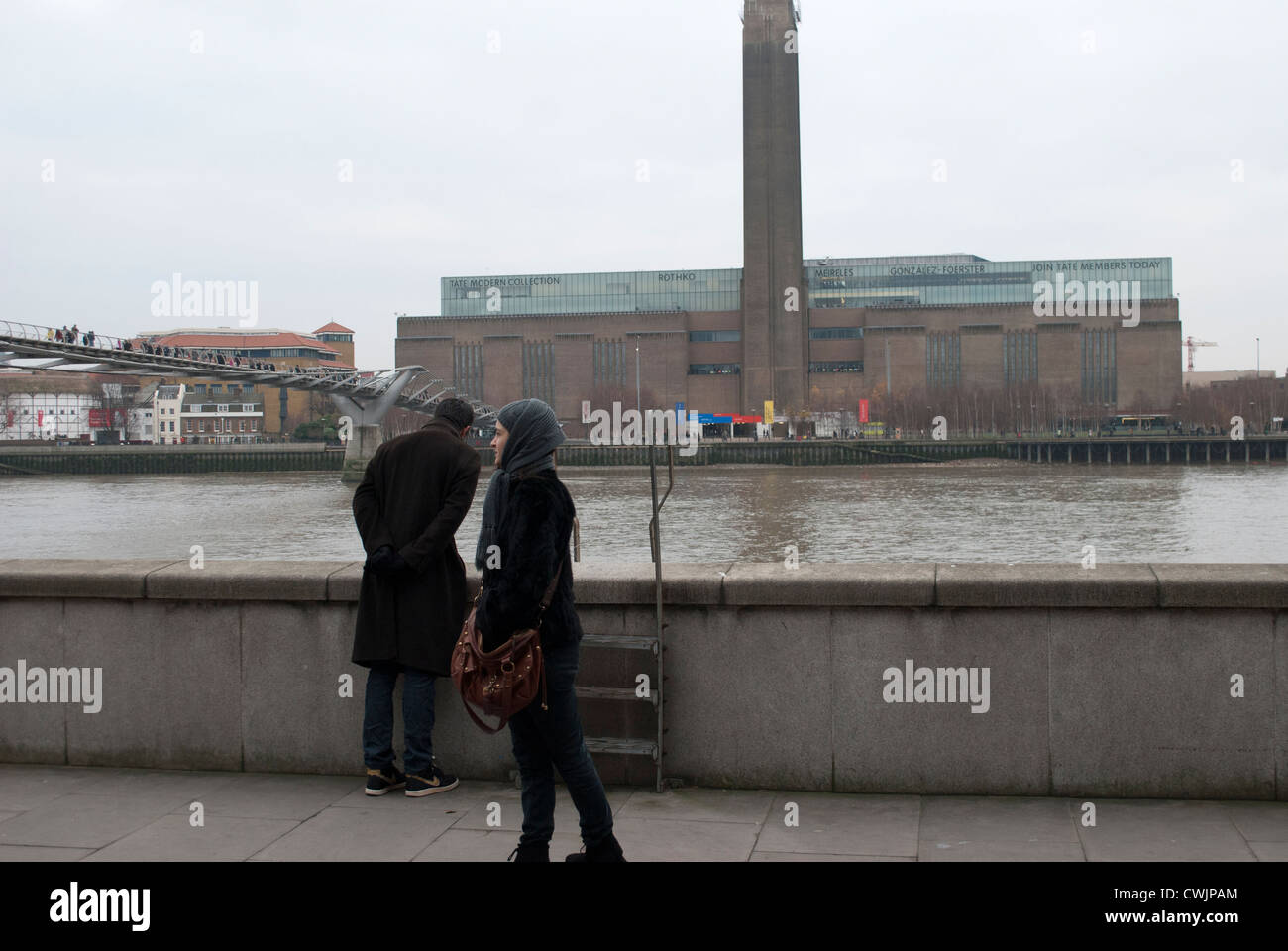 Tate Modern in the Bankside Power Station from the opposite bank of the Thames against a grey sky with two people Stock Photo