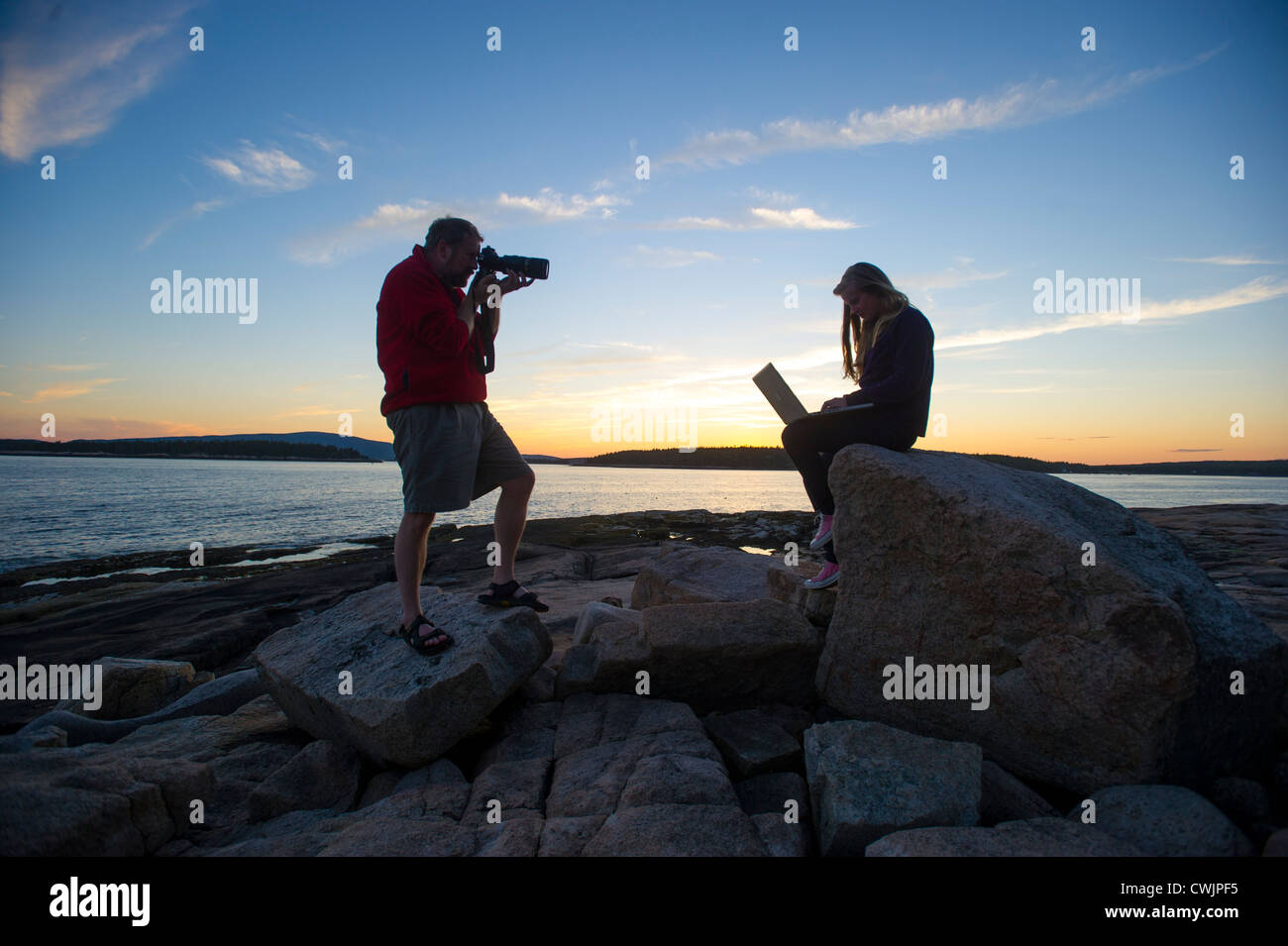 Photographer taking picture of woman using laptop at sunset, Acadia National Park, Maine USA Stock Photo