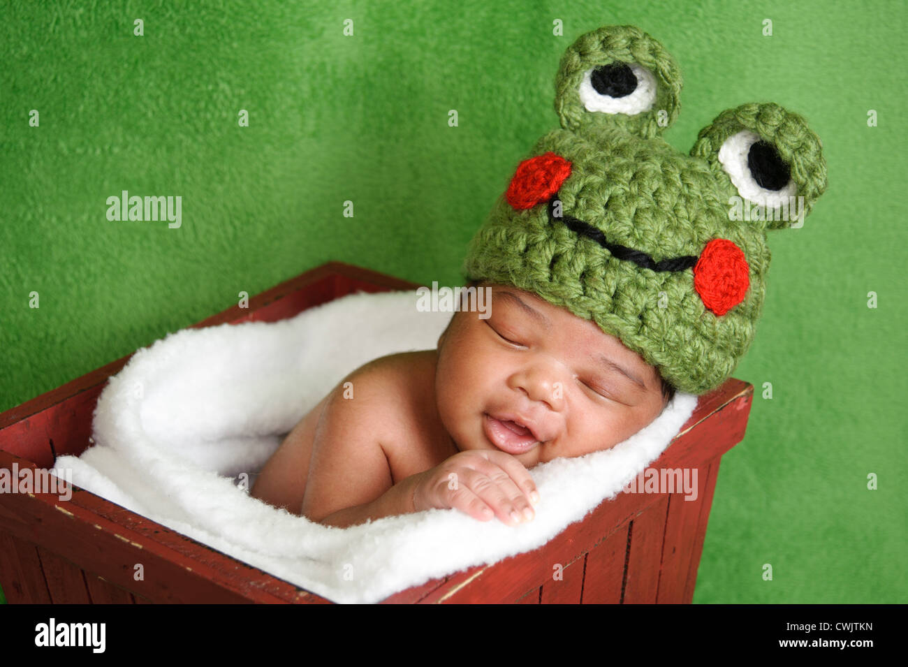 13 day old smiling newborn baby boy wearing a green crocheted frog hat. He is sleeping in a red wooden box. Stock Photo