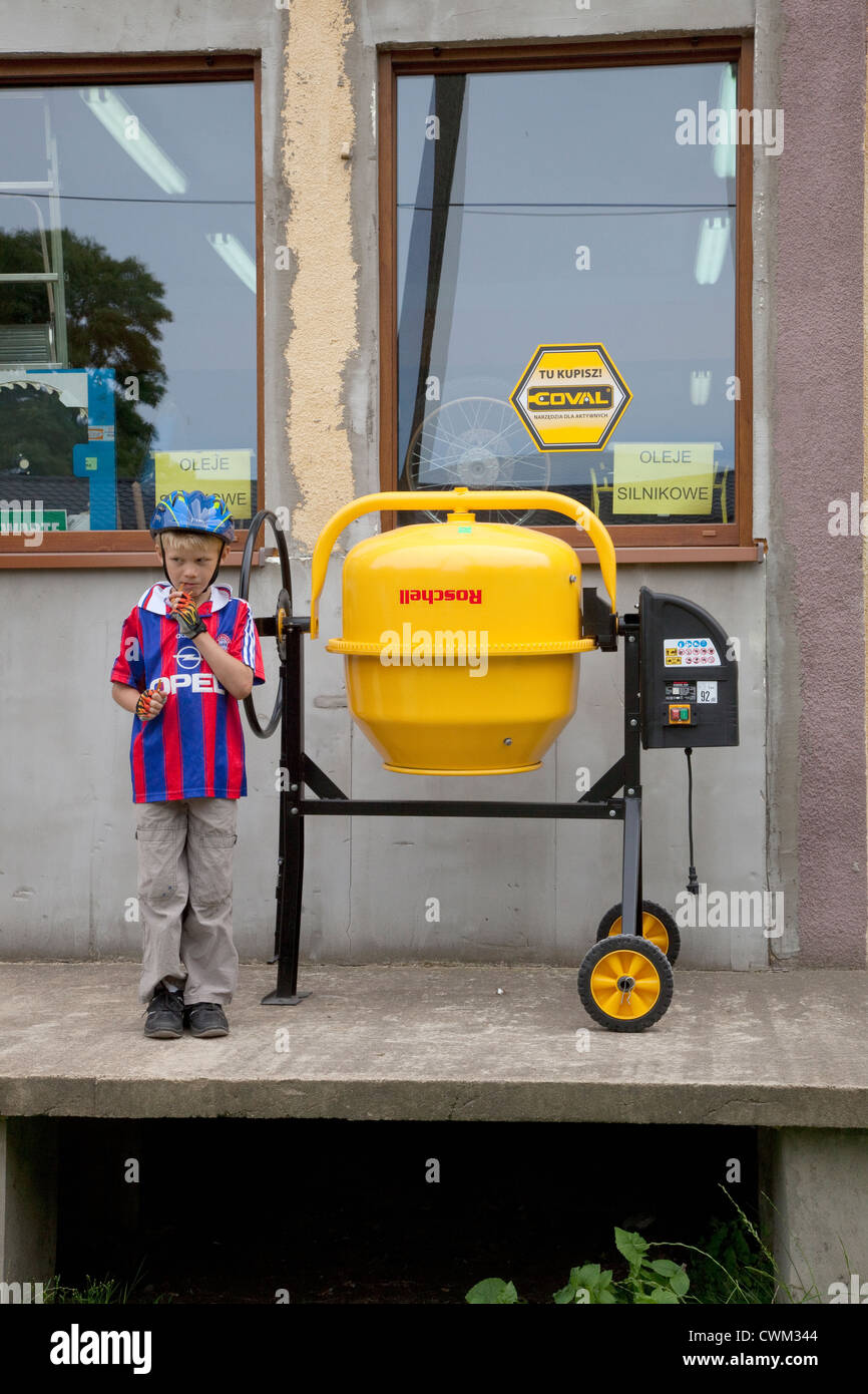 Polish boy age 8 standing beside yellow cement mixer in front of a hardware store. Rzeczyca Central Poland Stock Photo