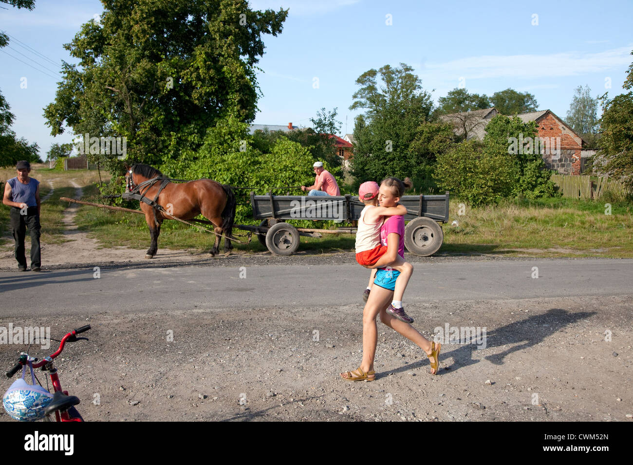 Teen girl carrying her brother along village street against a rural background of a horse drawn wagon. Mala Wola Central Poland Stock Photo