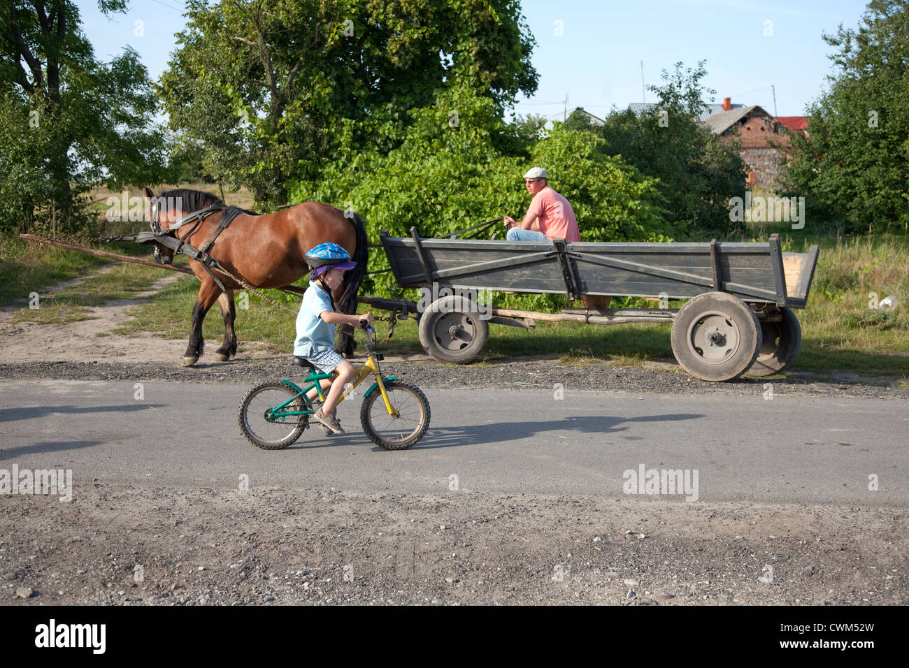 Helmeted Polish boy age 4 riding his bicycle down village lane beside horse drawn wagon. Mala Wola Central Poland Stock Photo