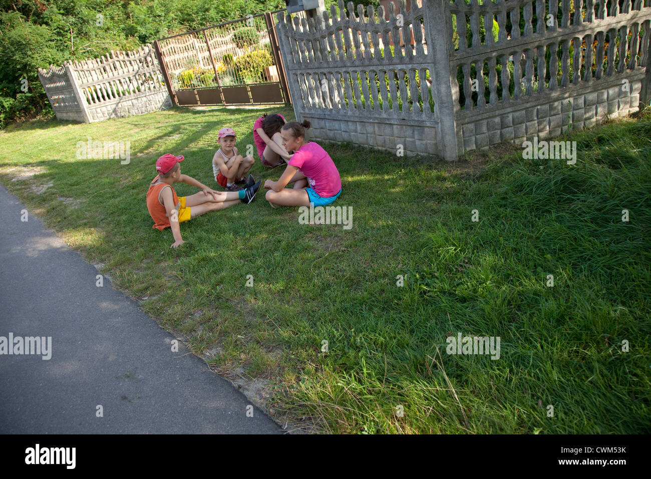 Polish village children and teens sitting on shaded green grass by fenced yard and their homes. Mala Wola Central Poland Stock Photo