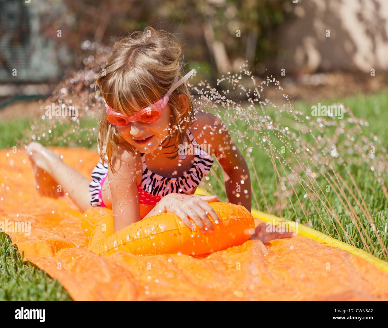 A little girl wearing goggles and bathing suit sliding on a slip and slide Stock Photo