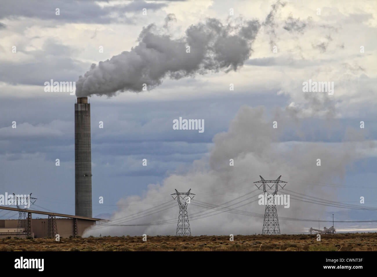 A smokestack spewing out pollutants while a power plant produces electricity.  Power lines and poles surround the plant. Stock Photo
