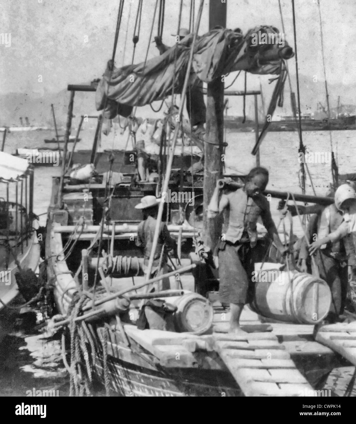 Unloading a Junk, Hong Kong, Two Chinese unloading a barrel, suspended from a pole over their shoulders, from a boat, circa 1900 Stock Photo