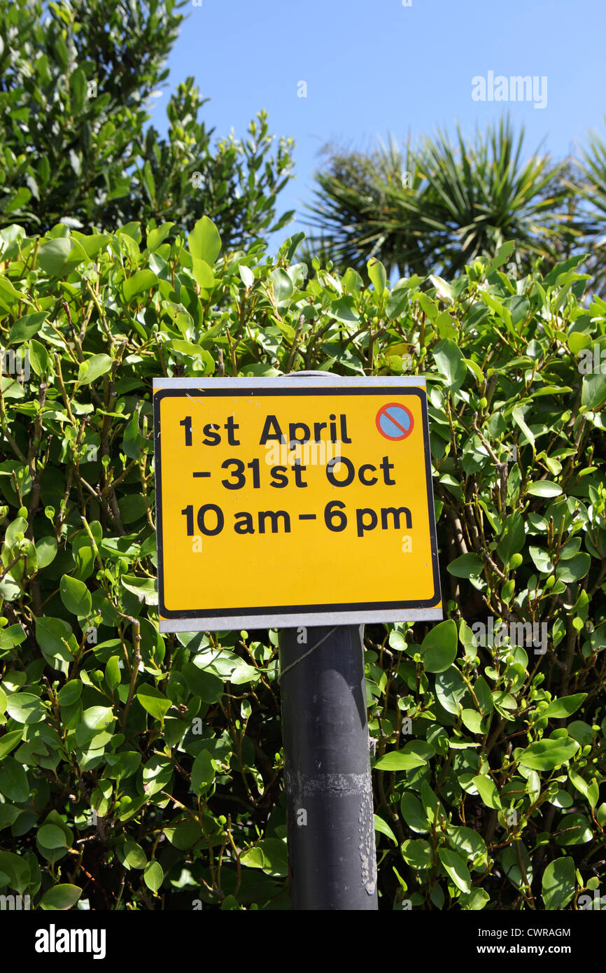 1st April - 31st October seasonal daytime parking restriction. Close-up photo of yellow sign against laurel hedge and blue sky Stock Photo