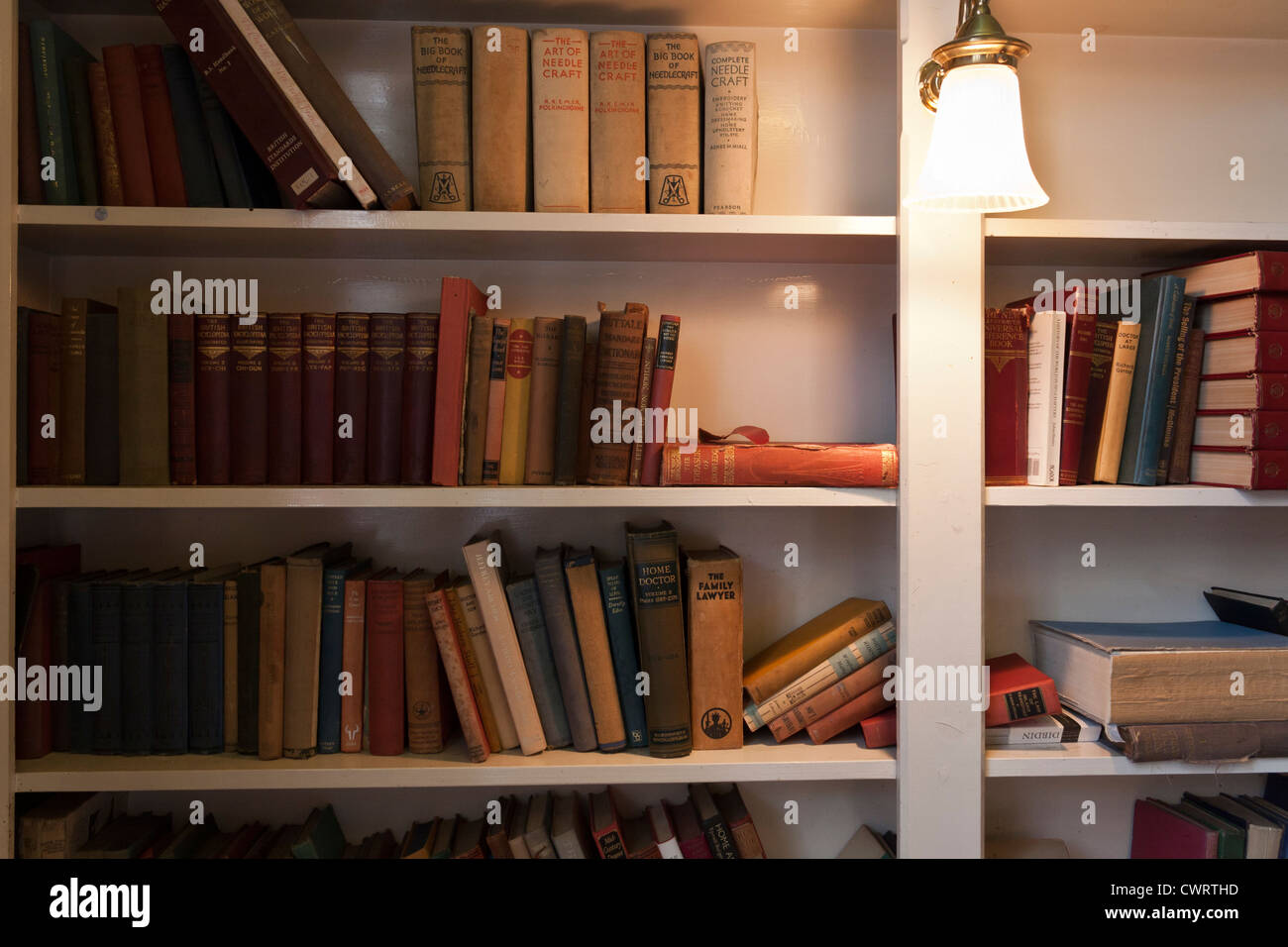 A variety of old well-worn books on a bookshelf, lit by a mounted lamp. Stock Photo