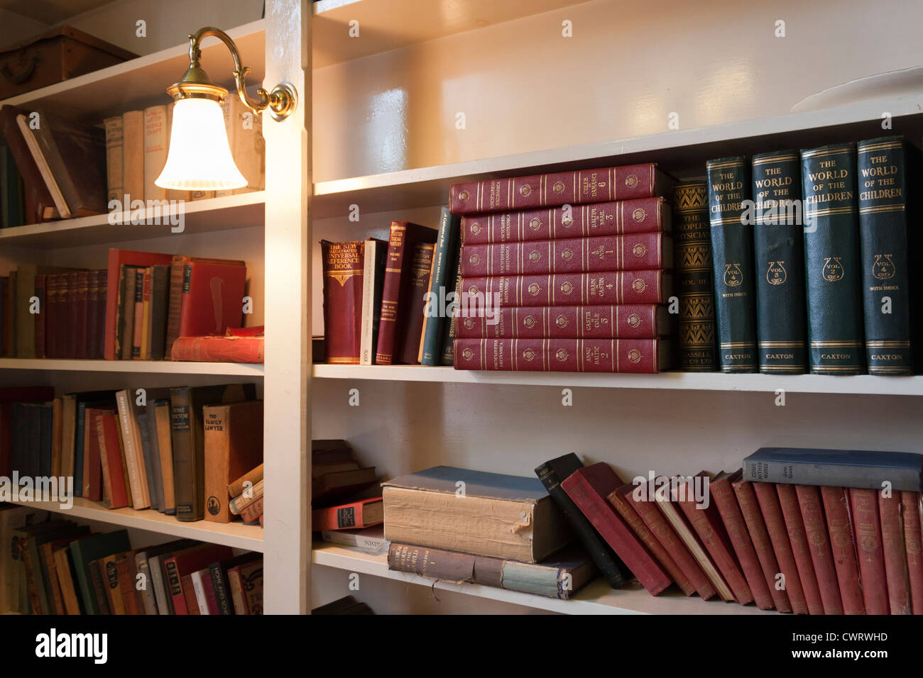 A variety of old well-worn books on a bookshelf, lit by a mounted lamp. Stock Photo