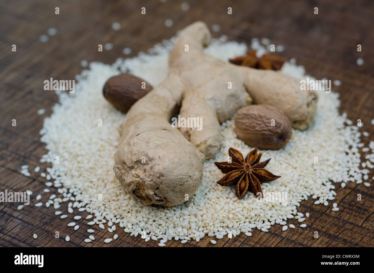 Spices, ginger root, star anise and nutmeg, arranged on a bed of hulled sesame seeds on a dark wooden surface. Stock Photo
