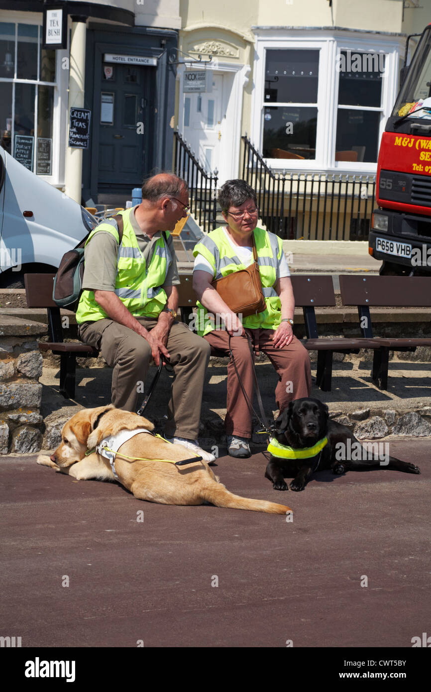 Visually impaired man and woman sat on bench enjoying the sunshine with guide dogs laid on the ground at their feet at Weymouth Stock Photo