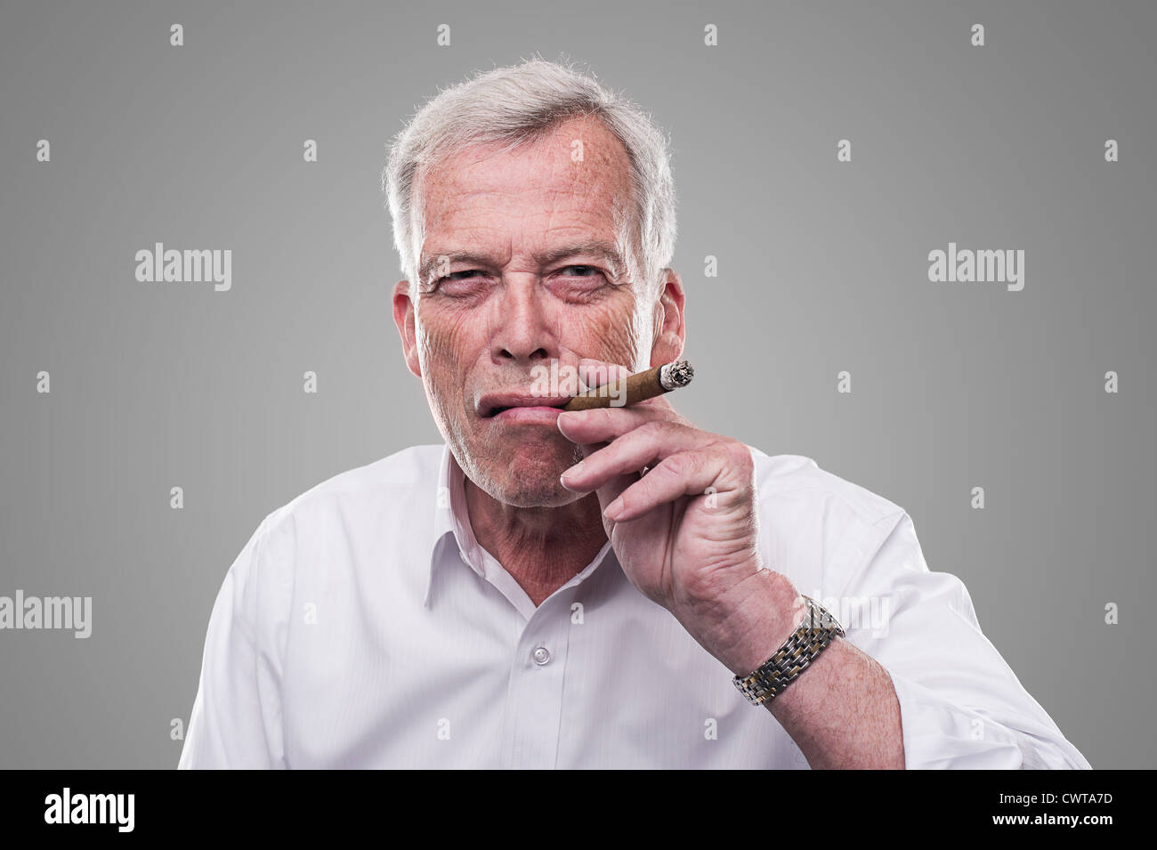 Handsome senior man with a shrewd expression and strong personaity smoking a cigar isolated on a grey studio background Stock Photo
