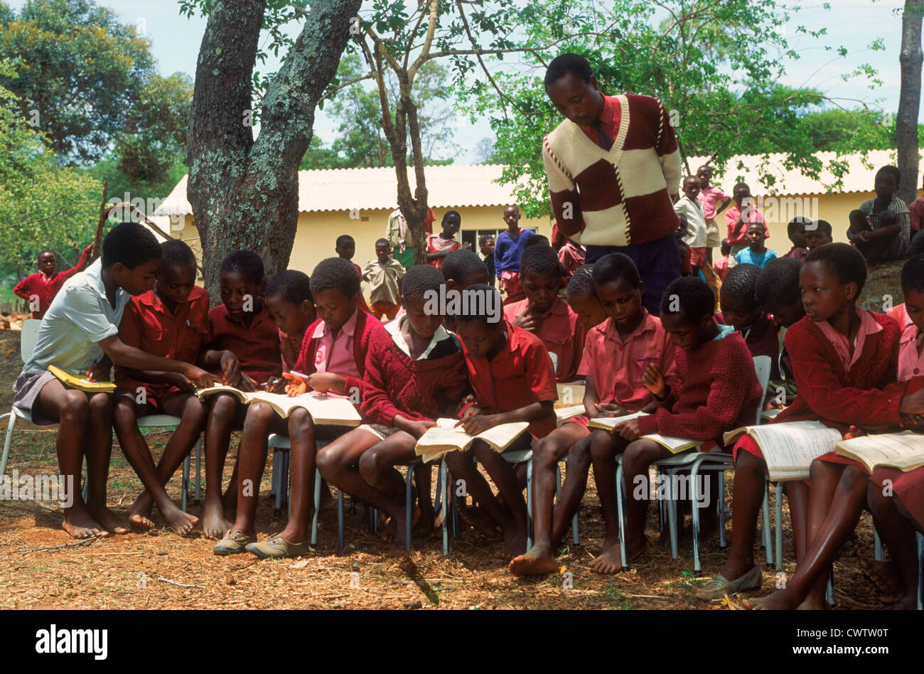 Rural school students 7 to 16 years with books  sitting in chairs outside in school yard studying with teacher in Zimbabwe Stock Photo