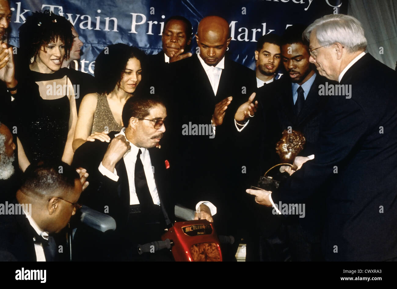 Comedian Richard Pryor, seated, receives the Mark Twain Comedy Award as family members look on at the Kennedy Center October 20, 1998 in Washington, DC (L-R) Daughters Rain Pryor, Elizabeth Pryor, actor Danny Glover, comedians Damon Wayans and Chris Rock. Stock Photo