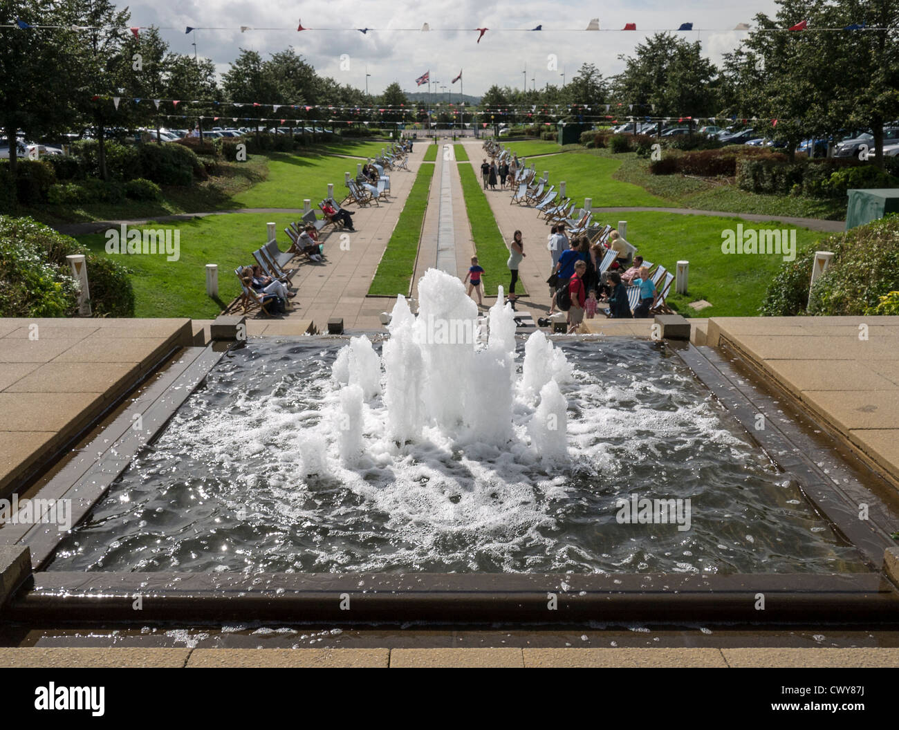 Shoppers relaxing in deck chairs by a water feature in the car park of