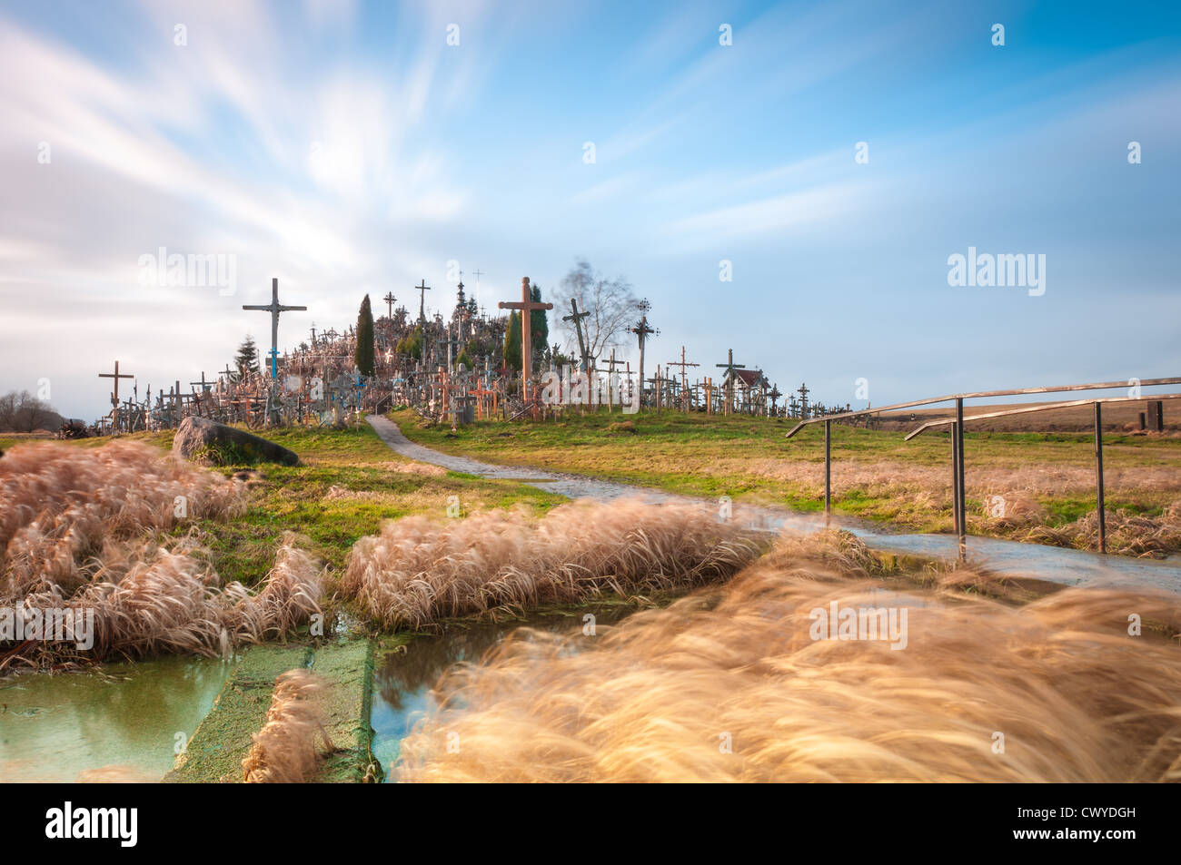 Famous hill of crosses near Siauliai, Lithuania, Europe. Yellow grass in foreground and blue cloudy sky in background. Stock Photo