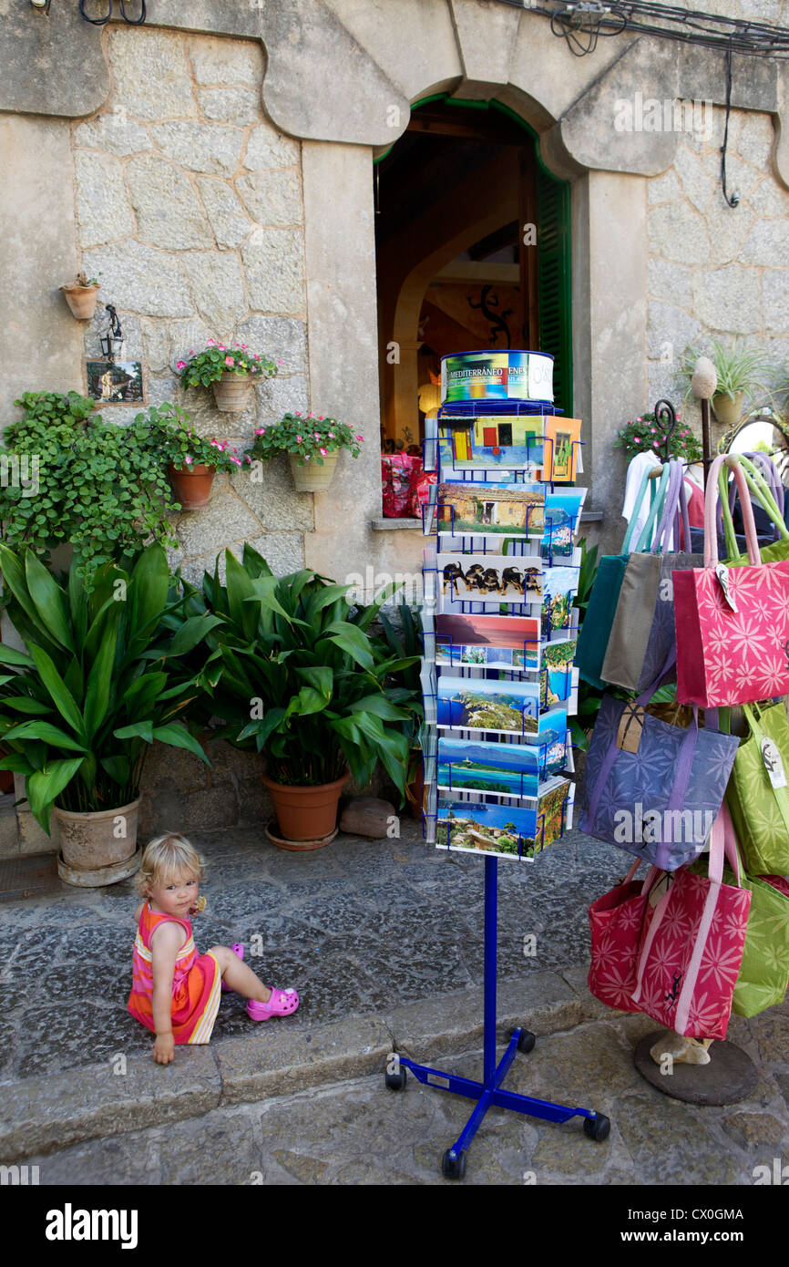 Little girl in a red dress sits on the pavement outside a shop beside a postcard stand and beach bag stand. Spain Mallorca Stock Photo