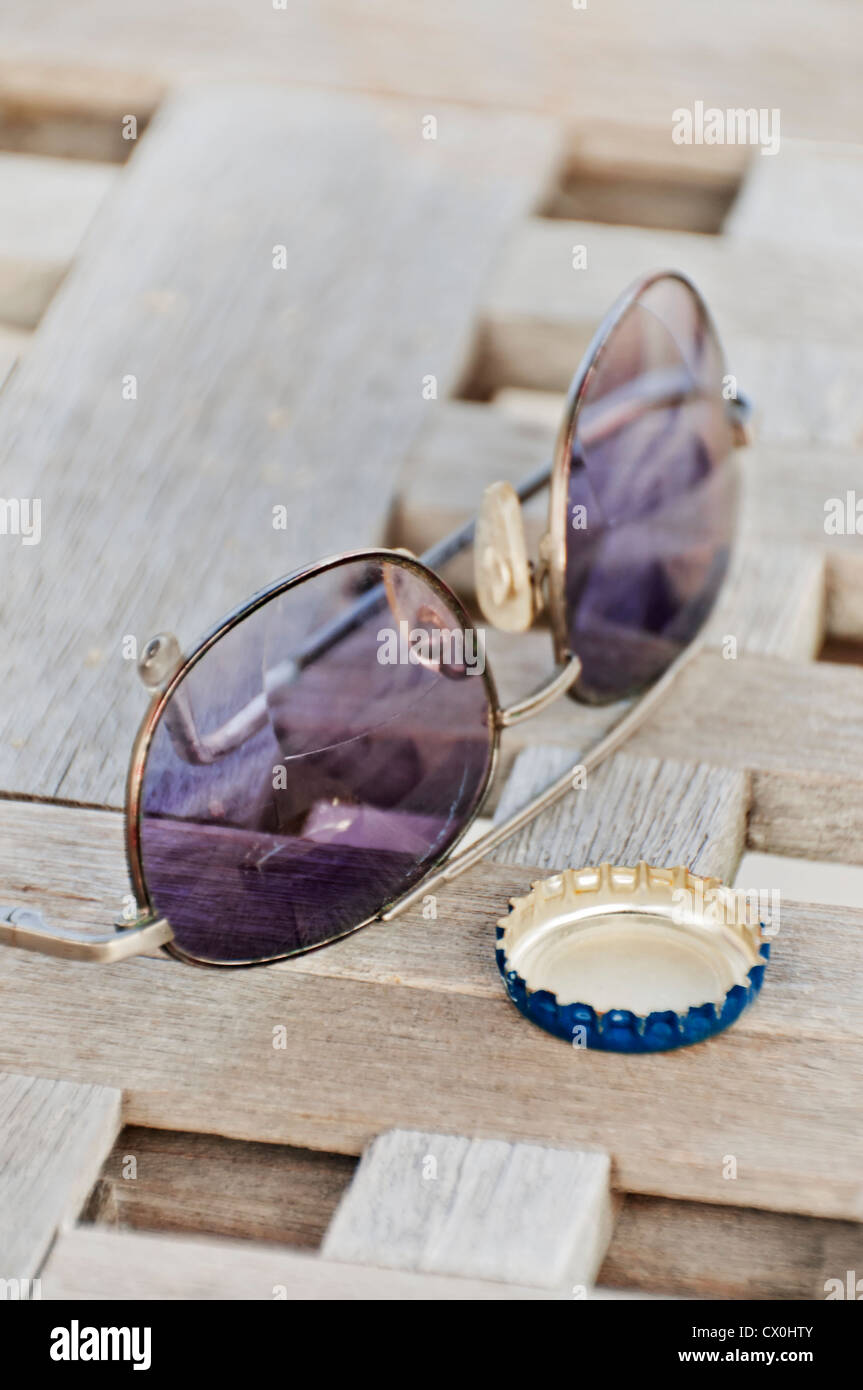 A pair of tinted sunglasses and a bottle cap are on a tabletop outdoors. Stock Photo