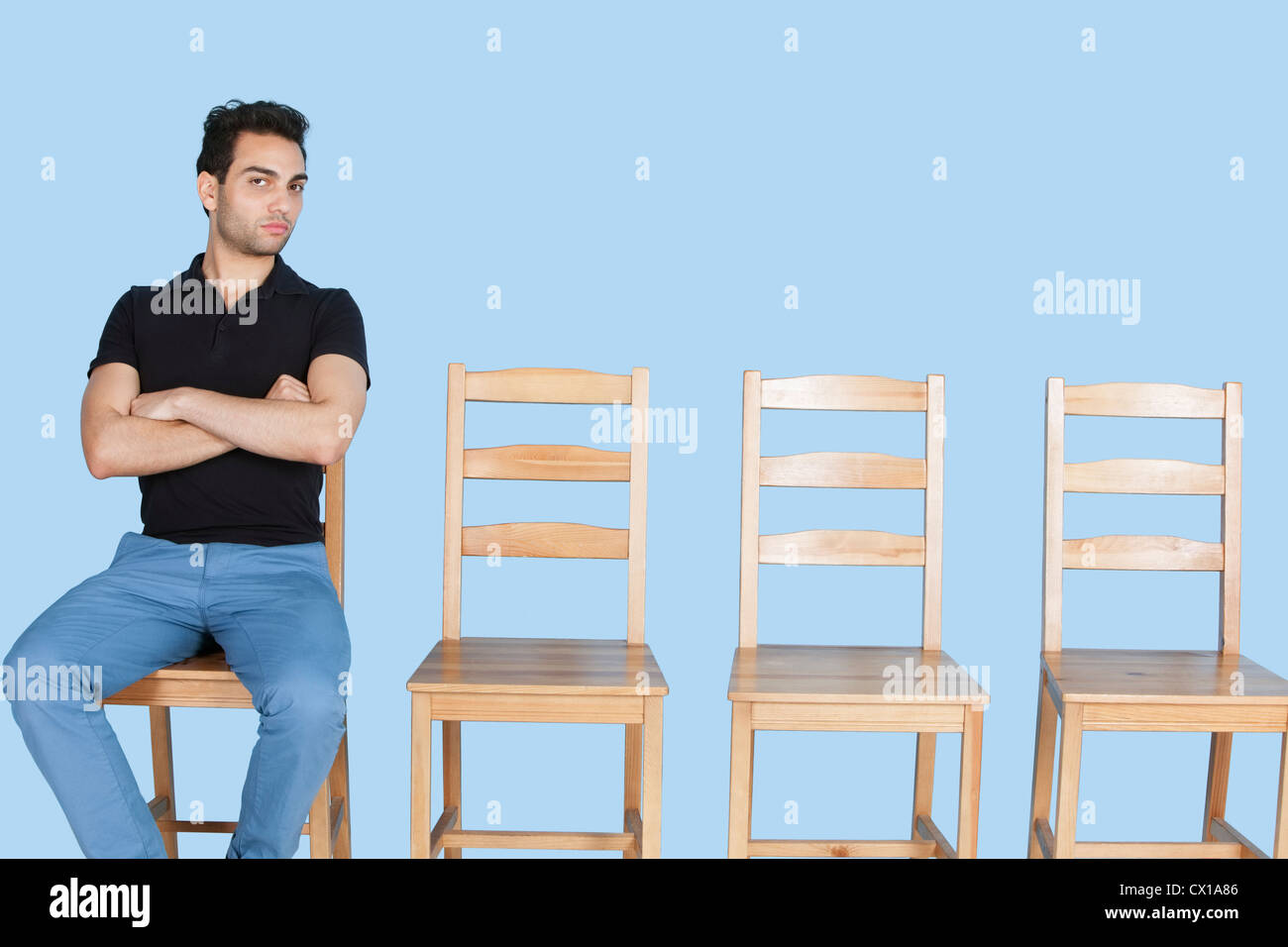 Portrait of a young man sitting beside empty wooden chairs over blue background Stock Photo