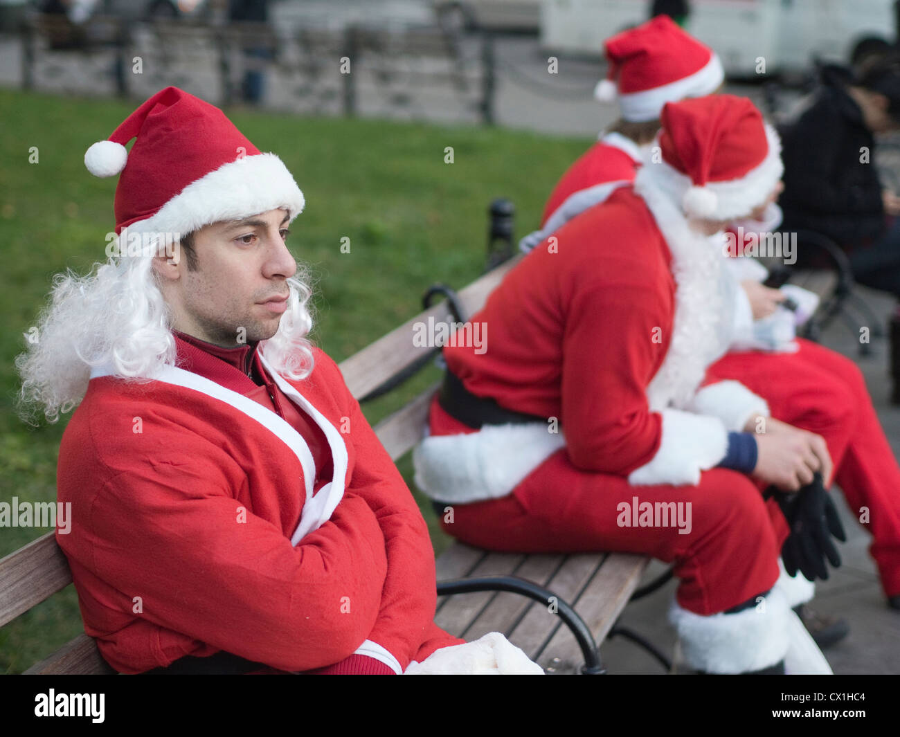 A Santa Claus Sits On A Park Bench With Other Santas During Santa
