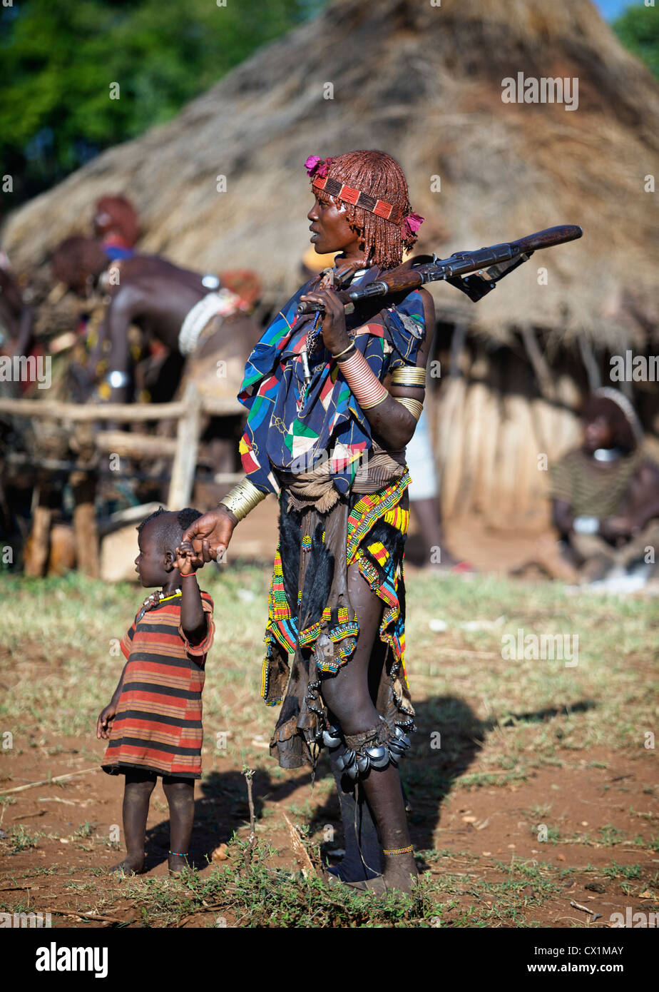 Hamar woman holding an AK47 while looking after her child during a Bull Jumping ceremony. Stock Photo