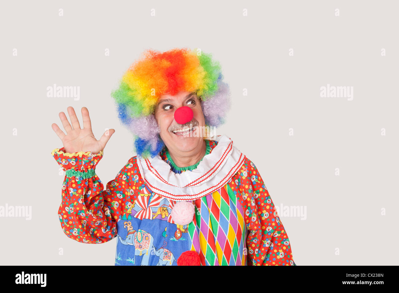 Cheerful senior male clown waving hand while looking away over gray background Stock Photo
