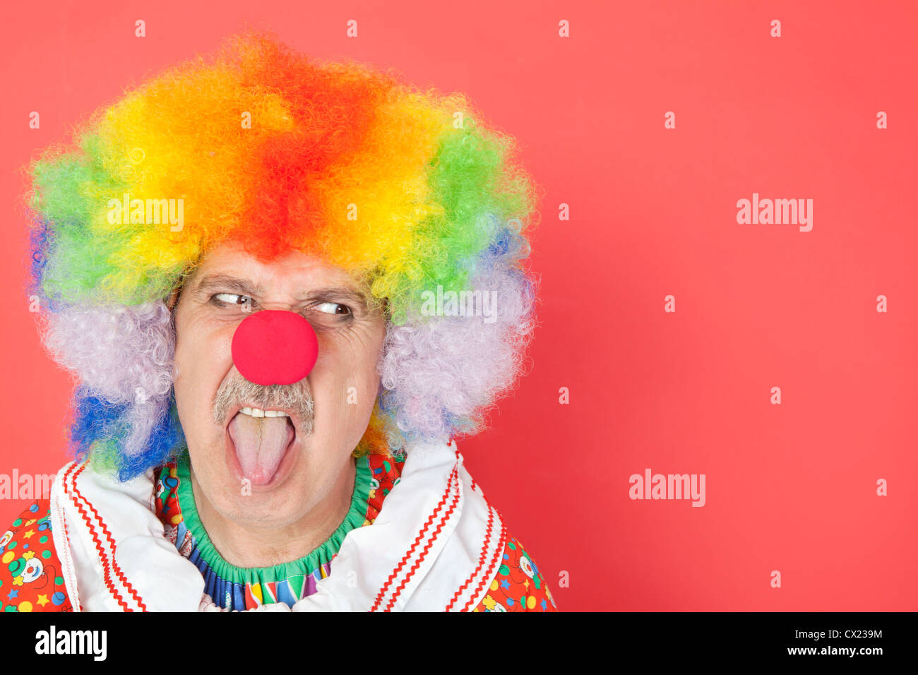 Senior male clown sticking out tongue while looking away over red background Stock Photo