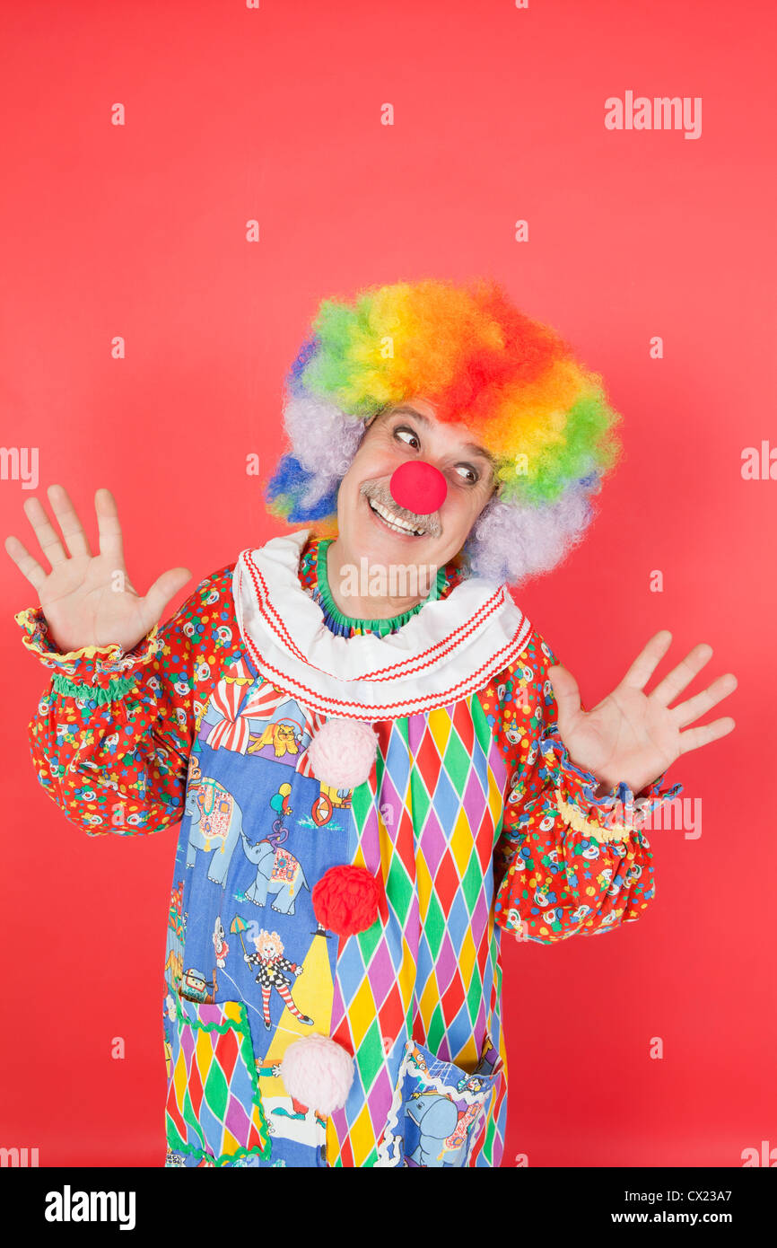 Funny clown with arms raised looking away against colored background Stock Photo