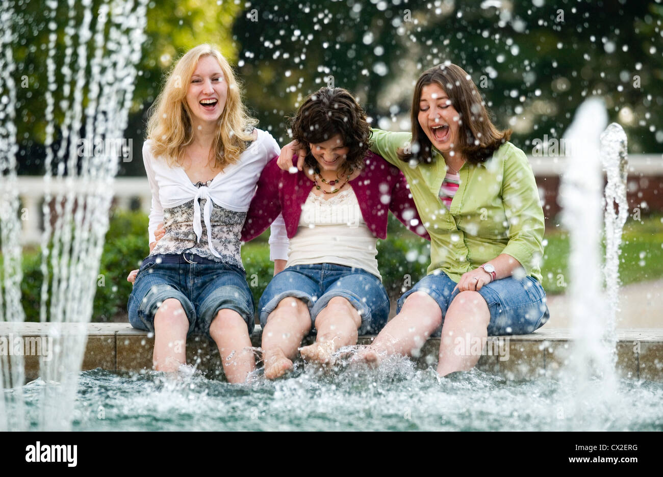 Three girls sit on the edge of a fountain laughing as they dip their feet in the water. Stock Photo