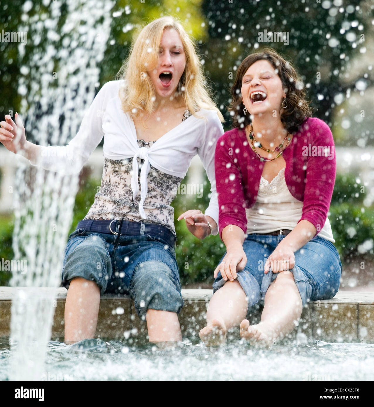 Two girls sit on the edge of a fountain laughing as they dip their feet in the water. Stock Photo