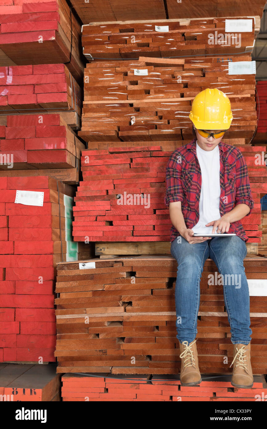 Asian female industrial worker using tablet PC while sitting on stacked wooden planks Stock Photo