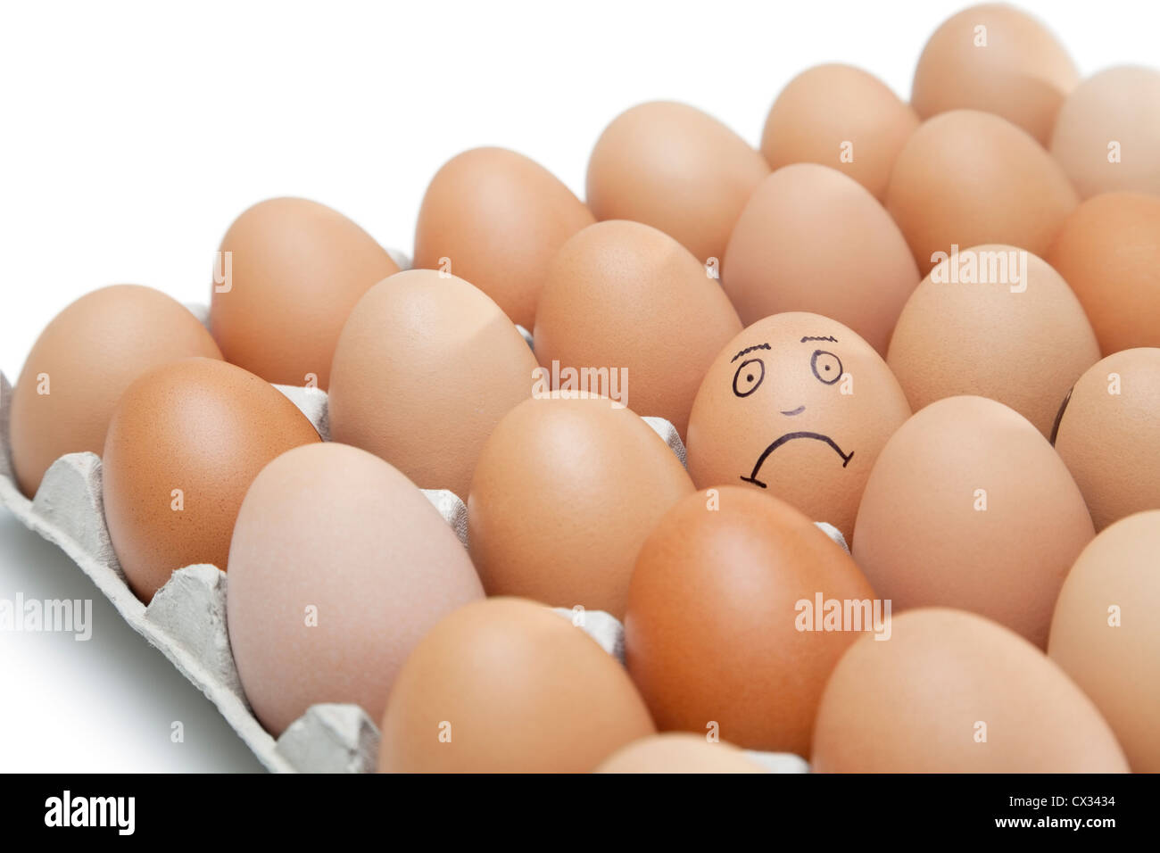 Sad face drawn on an egg surrounded by plain brown eggs in carton against white background Stock Photo