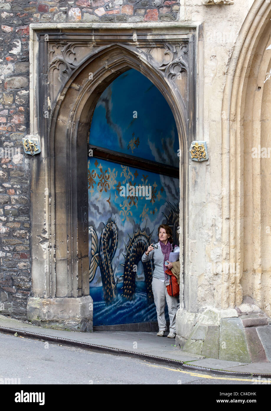 A woman with a camera standing in front of a mural depicting fantastical and mythical creatures at St. John's Gate in Bristol Stock Photo