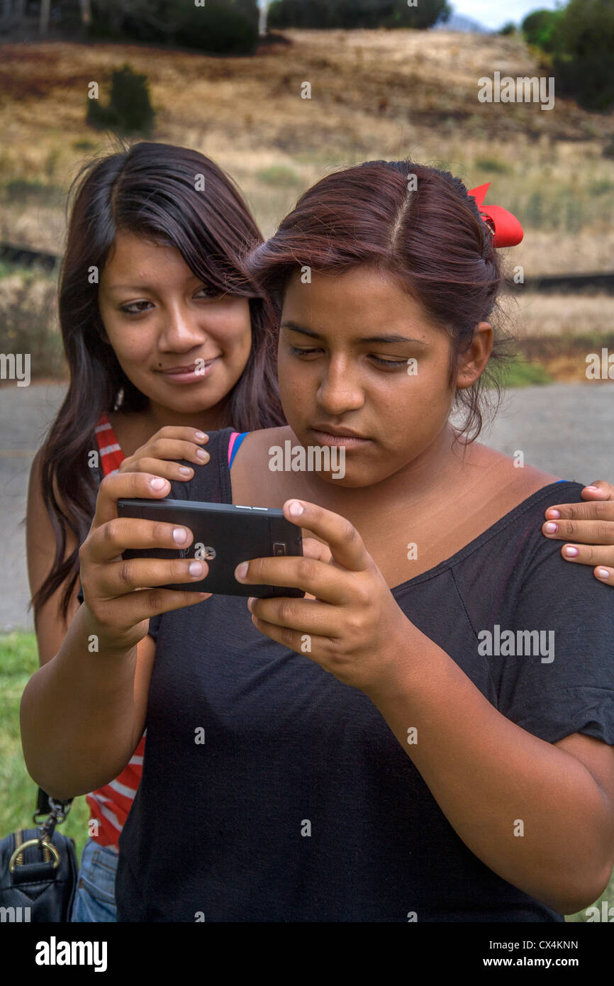An upset Hispanic girl reads a bullying message on her Smart Phone as her friend comforts her. Stock Photo