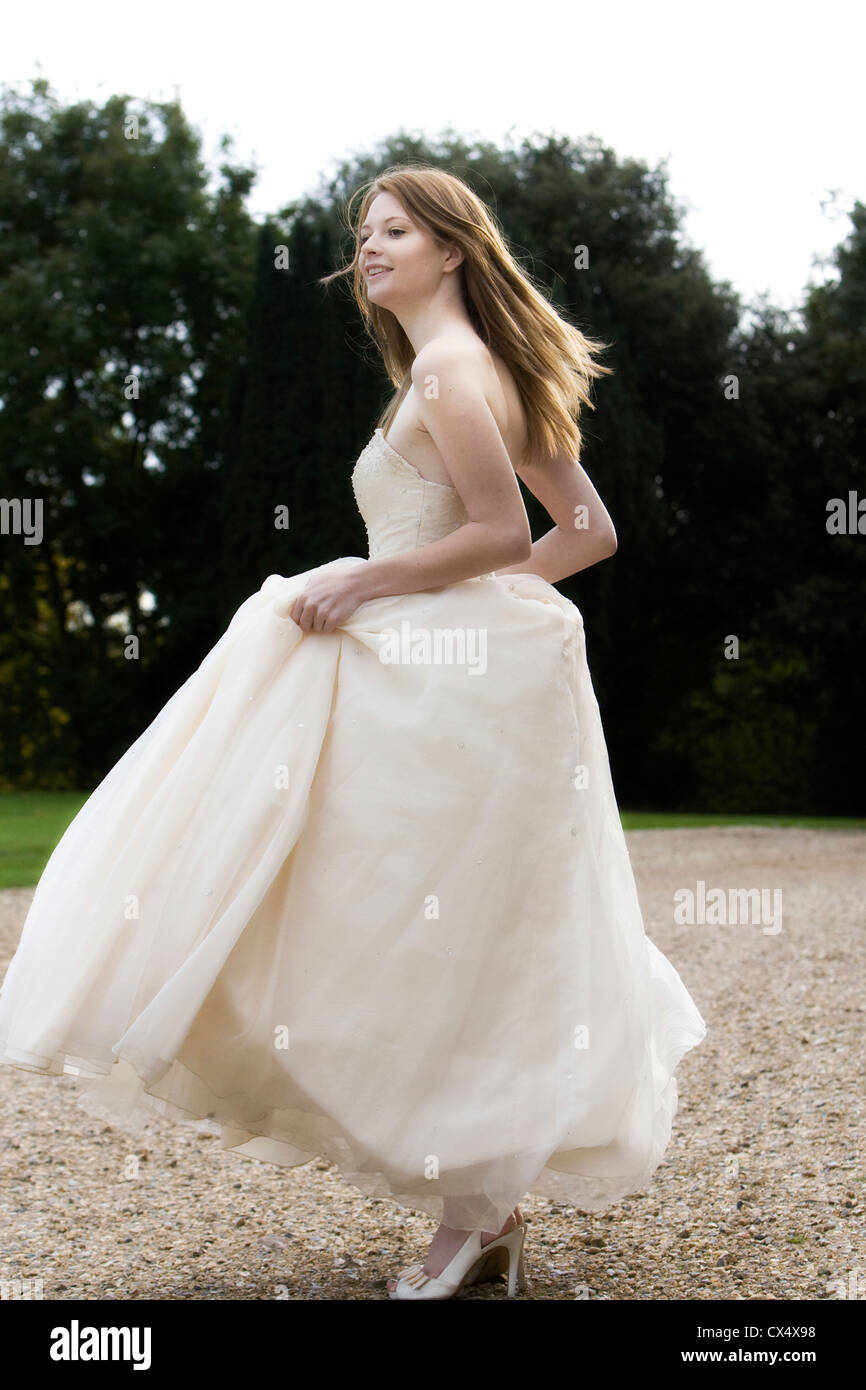 Side view of young woman in cream bridal dress holding skirts in grounds of stately home Stock Photo